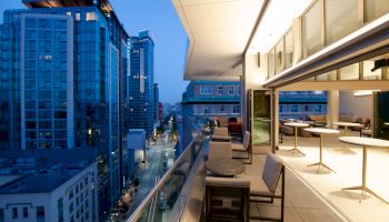 The image shows a modern rooftop terrace with seating, overlooking a cityscape of tall buildings at dusk.