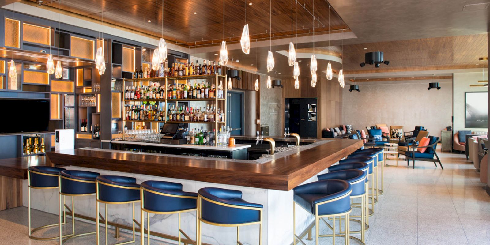 The image shows a modern bar interior with a central island, stylish bar stools, hanging lights, and a well-stocked liquor shelf.