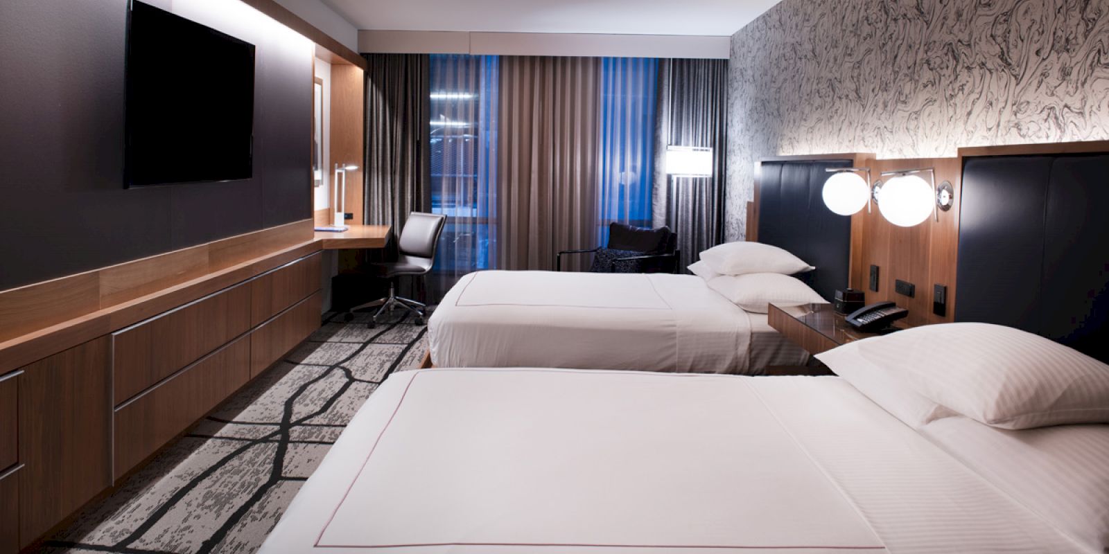 The image shows a modern hotel room with two double beds, a large TV, a work desk with a chair, and stylish lighting and decor.