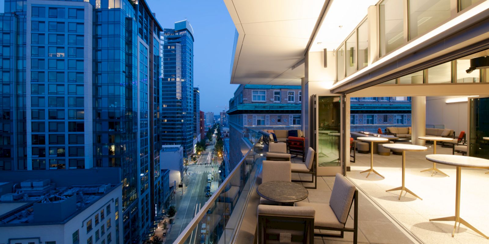 A modern rooftop patio with tables and chairs overlooks a cityscape of tall buildings and a street at dusk.