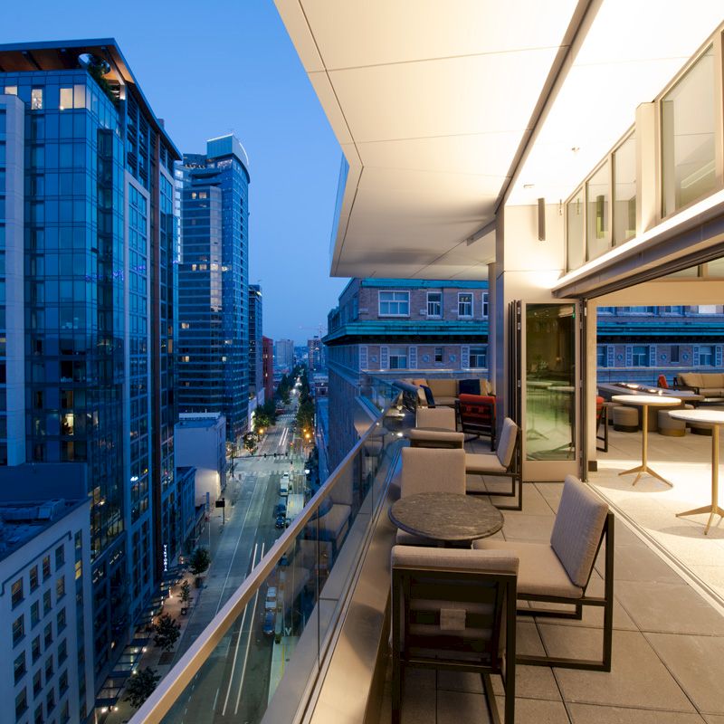 A modern rooftop terrace overlooking an urban street, featuring tables, chairs, and city buildings at dusk with ample glass and lighting.