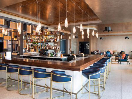 A modern bar features hanging lights, blue stools, a well-stocked bar, and a seating area with couches and chairs in the background.