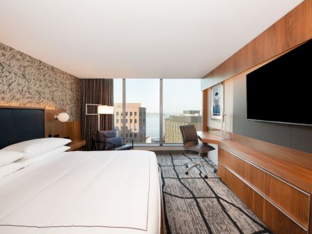 A modern hotel room with a large bed, desk, TV, and window with a city view.