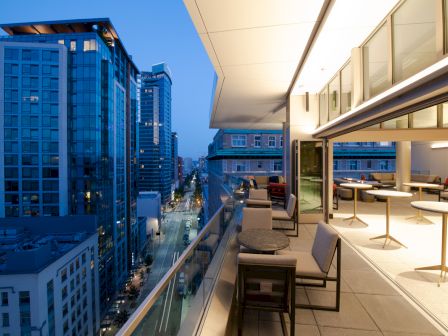 Modern urban balcony with tables and chairs, overlooking a city street and tall glass buildings at dusk.