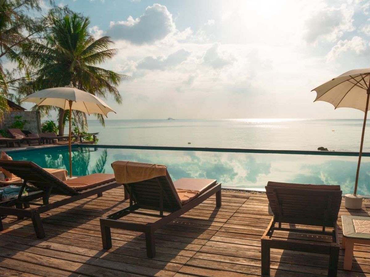 Poolside scene with loungers, umbrellas, palm trees, and a tranquil ocean view under a cloudy sky.