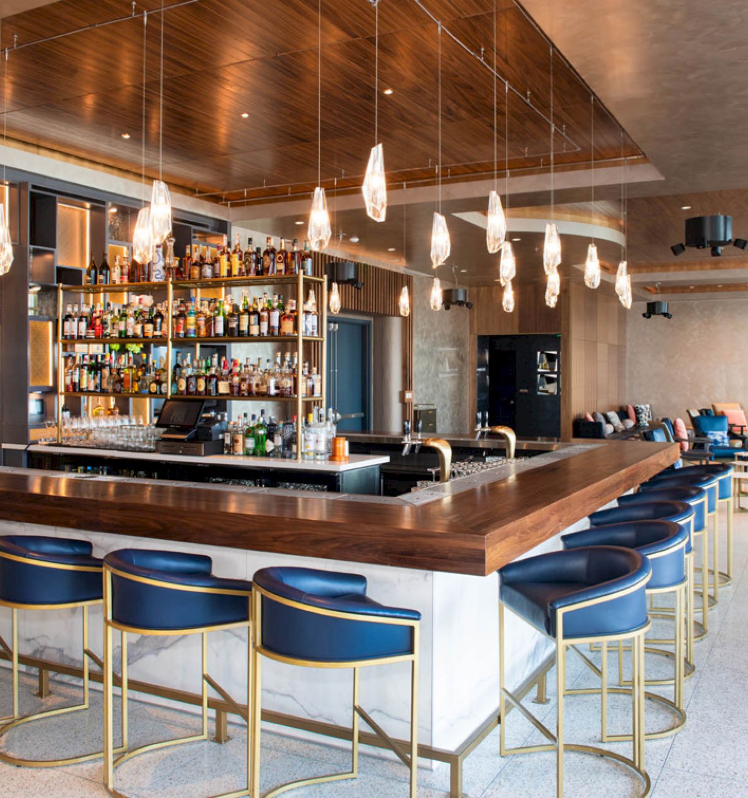 The image shows a modern bar with blue stools, a wooden countertop, and hanging lights, surrounded by shelves with bottles and lounge seating.