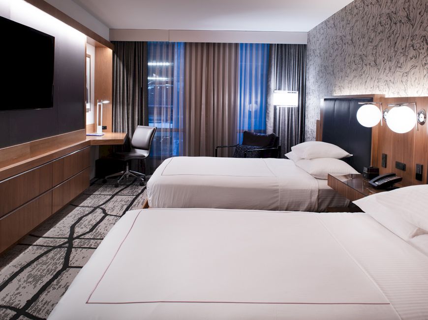 The image shows a modern hotel room with two beds, a desk, a TV on the wall, and patterned curtains and wallpaper.