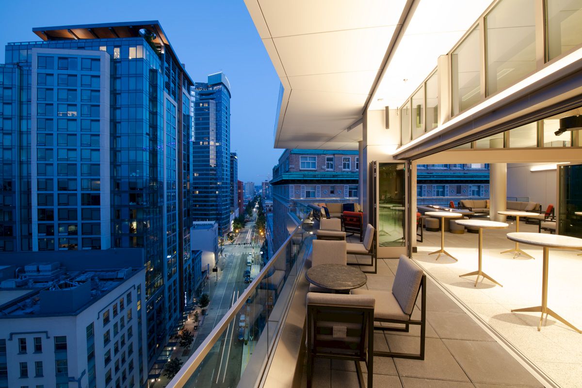 A modern balcony view at dusk overlooking a city street with tall buildings. Tables and chairs are arranged on the balcony.