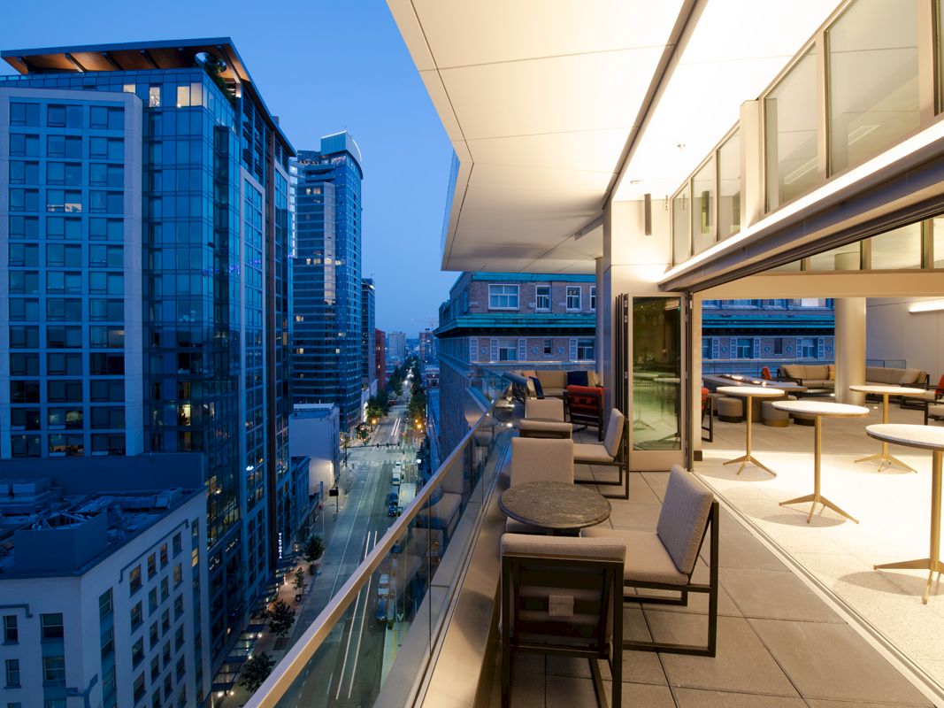 A modern balcony view at dusk overlooking a city street with tall buildings. Tables and chairs are arranged on the balcony.