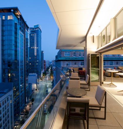 A modern rooftop terrace overlooking city buildings at dusk. The terrace has seating and tables with a sleek, open design.