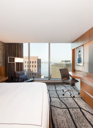 A modern hotel room with a bed, desk, large TV, and a window view of buildings and water in the background.