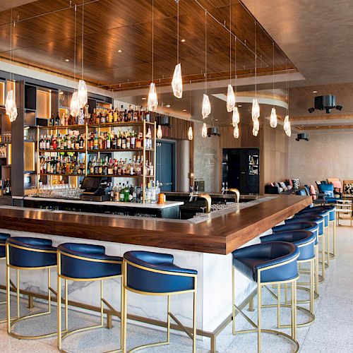 A modern bar with blue stools, wooden accents, a stocked liquor shelf, and ambient lighting, set in a spacious lounge area.