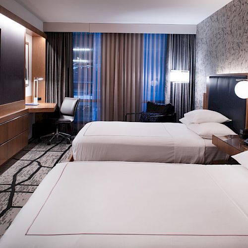 A modern hotel room with two beds, a wall-mounted TV, desk, and chair. Lamps provide cozy lighting next to the beds.