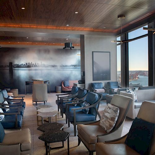 A modern lounge with plush seating overlooks a waterfront view, featuring large windows, warm lighting, and skyline artwork on the wall.