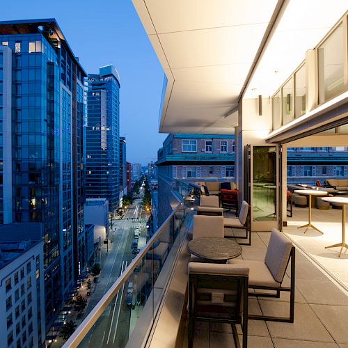 Rooftop terrace overlooking a city street at twilight, with tables and chairs, glass railings, and modern buildings in the background.