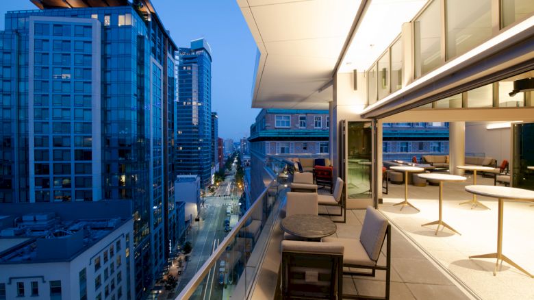 A modern cityscape at dusk with tall buildings and a stylish balcony featuring tables and chairs.