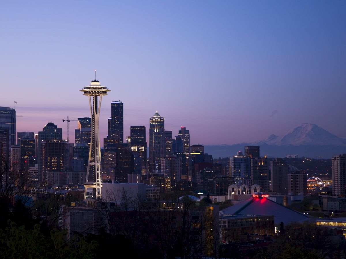Seattle skyline at dusk with the Space Needle and Mount Rainier visible in the background, under a clear sky.