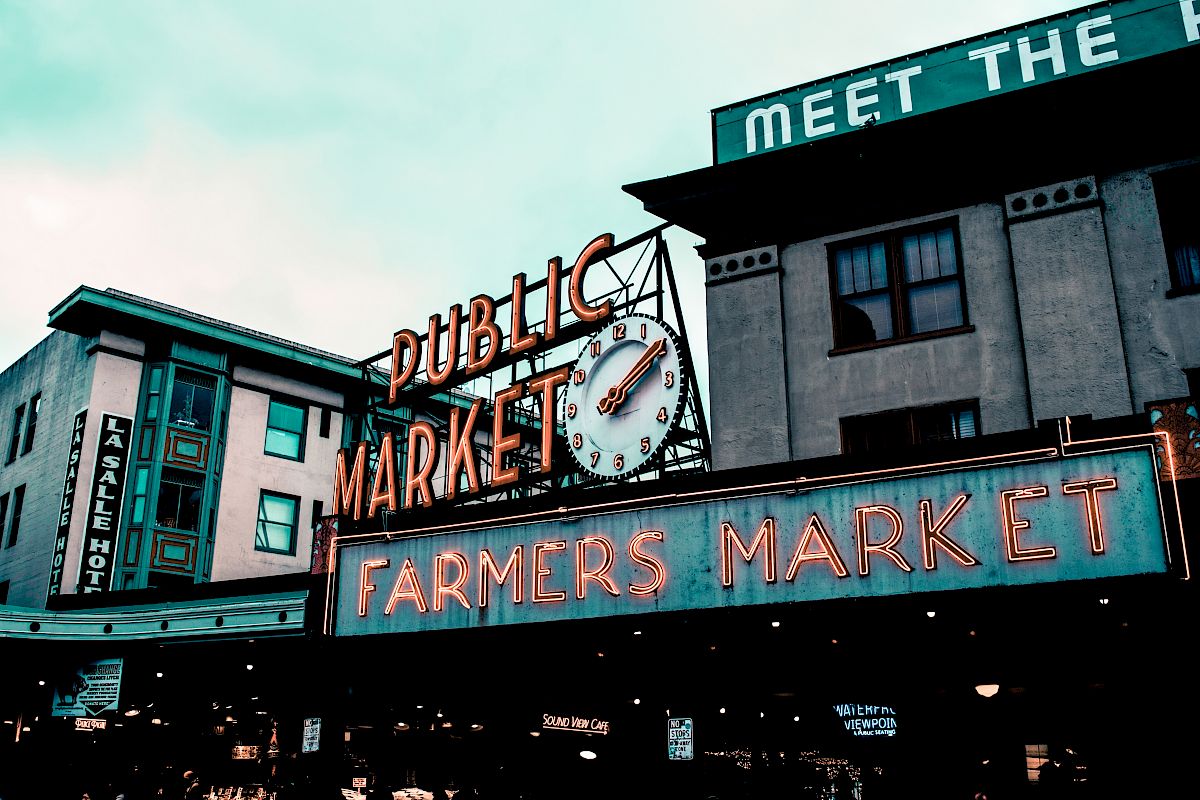 The image shows a classic neon sign for "Public Market" and "Farmers Market" with an iconic clock, likely located in an urban setting.