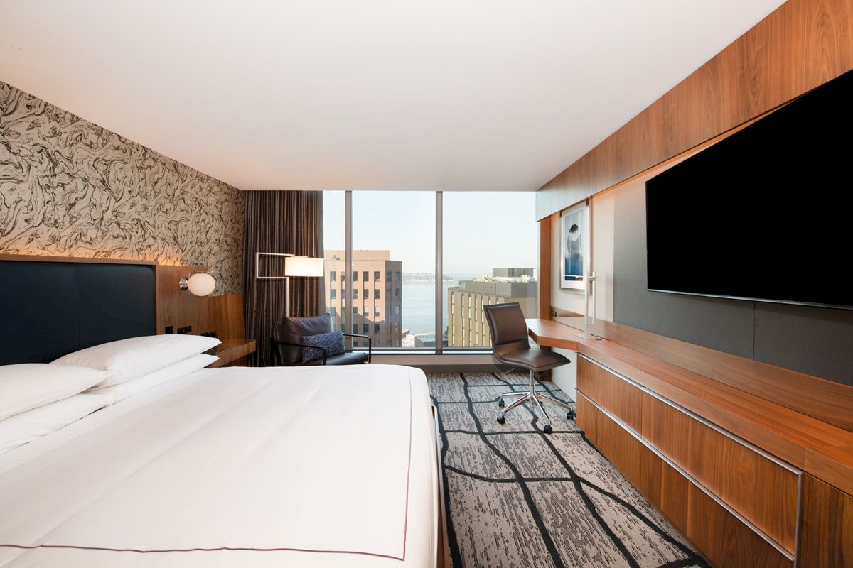 A modern hotel room with a large bed, desk, chair, TV, and a view of buildings through a wide window.