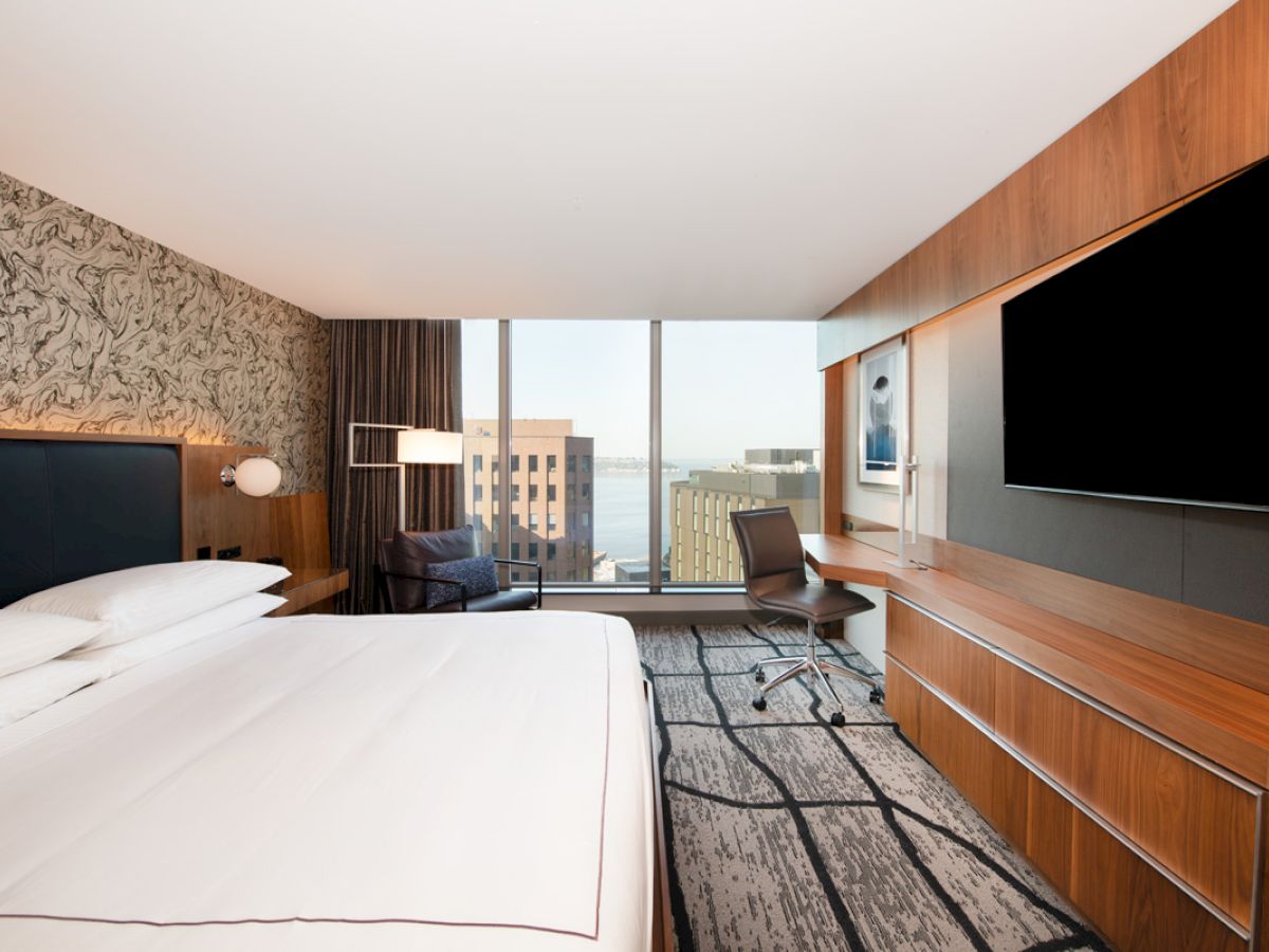 A modern hotel room with a large bed, desk, chair, TV, and a view of buildings through a wide window.
