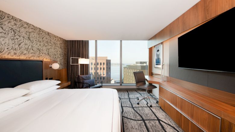 A modern hotel room with a large bed, desk, chair, TV, and a view of buildings through a wide window.