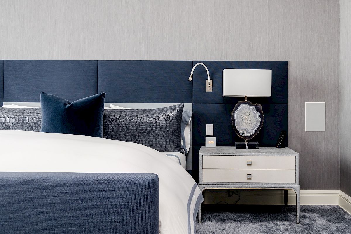 A modern bedroom with a blue and white color scheme, featuring a bed, pillows, nightstand, lamp, and decorative accessories.