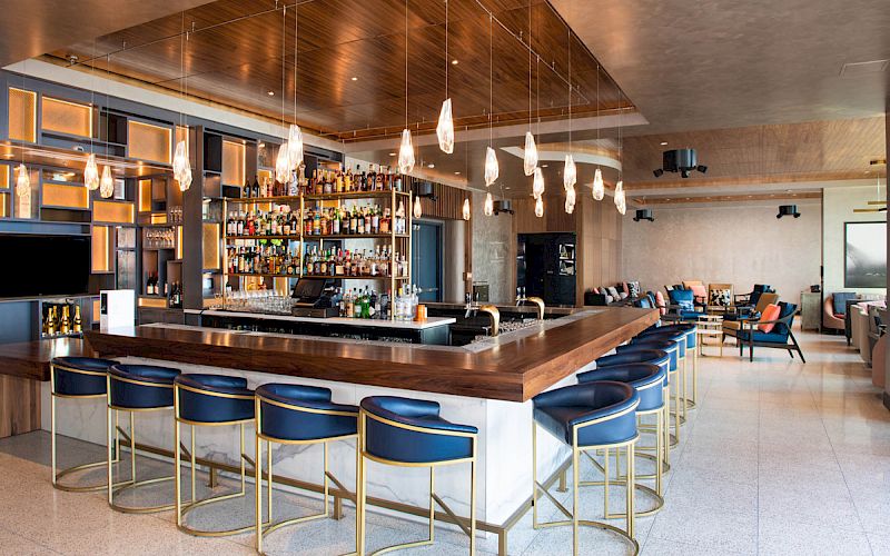 A modern bar with blue stools and pendant lights, featuring a well-stocked back bar and a lounge area with tables and chairs.
