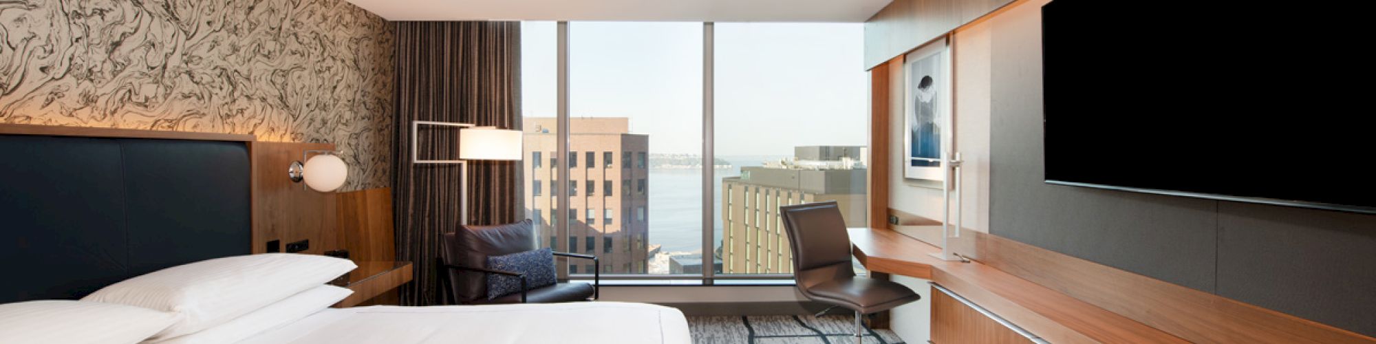 A modern hotel room with a large bed, wall-mounted TV, desk, chair, and a window with a city view.