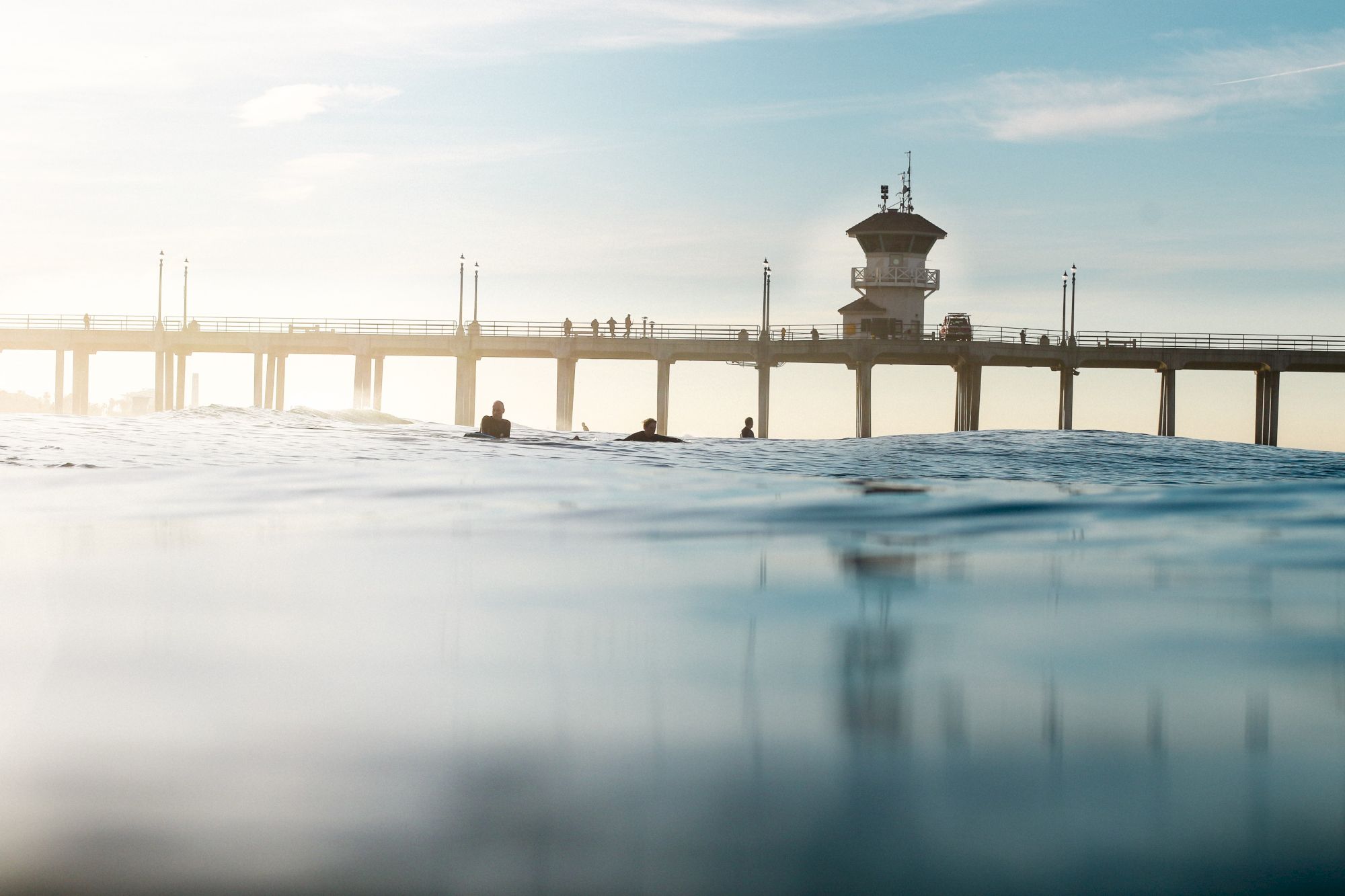The image shows a view of a pier extending into the ocean with a few people swimming nearby and a lifeguard tower in the background.